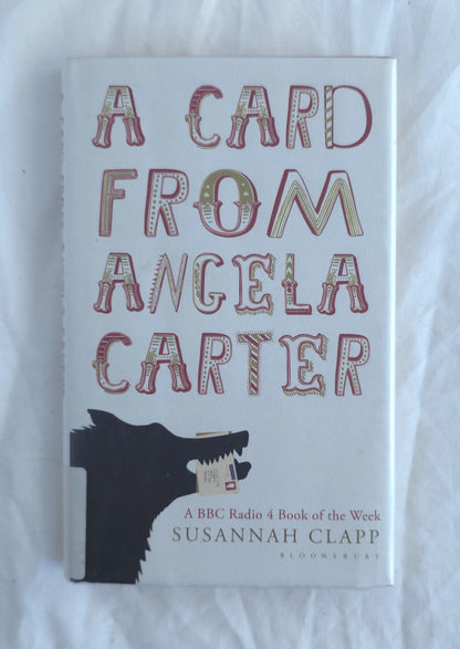 The book cover of 'A Card from Angela Carter' by Susannah Clapp, featuring a white background with stylized red and gold lettering and a black wolf illustration at the bottom, photographed on a white fabric surface.