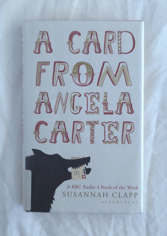 The book cover of 'A Card from Angela Carter' by Susannah Clapp, featuring a white background with stylized red and gold lettering and a black wolf illustration at the bottom, photographed on a white fabric surface.