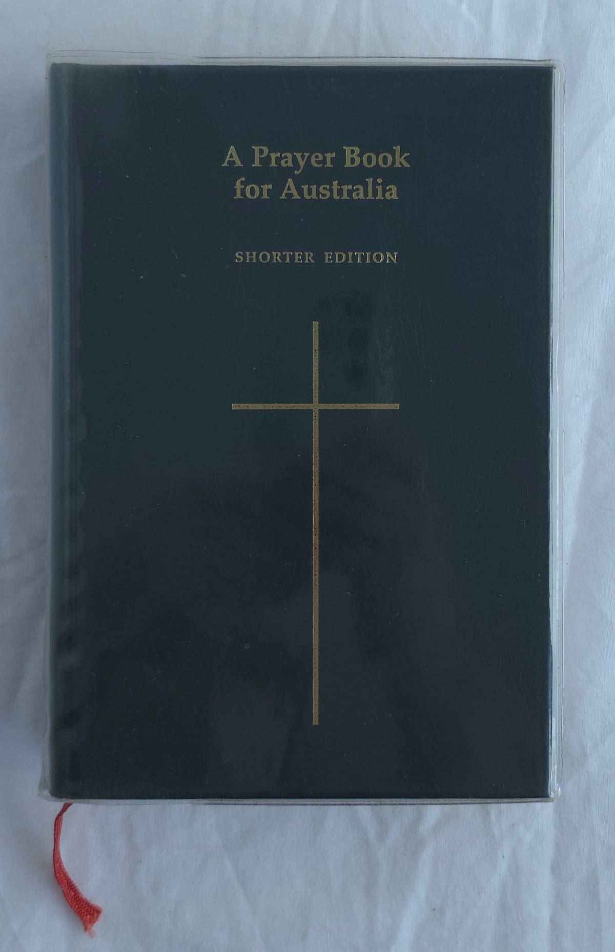 A Prayer Book for Australia Shorter Edition book with a black cover, gold cross and text, and a red ribbon bookmark, photographed on a white fabric background.