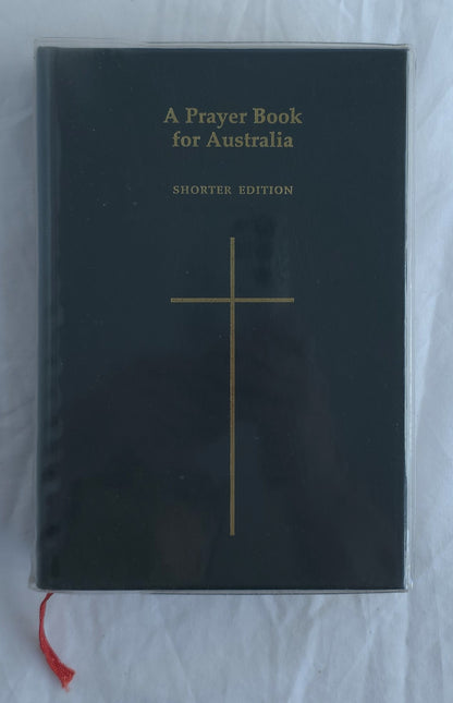 A Prayer Book for Australia Shorter Edition book with a black cover, gold cross and text, and a red ribbon bookmark, photographed on a white fabric background.