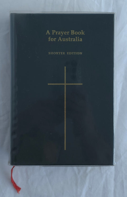 A Prayer Book for Australia Shorter Edition book with a black cover, gold cross and text, and a red ribbon bookmark, photographed on a white fabric background.