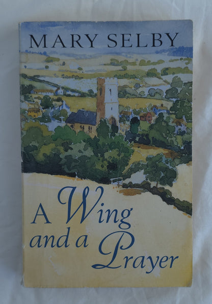 A vintage paperback book of Mary Selby's 'A Wing and a Prayer' resting on a white fabric surface. The cover features a watercolor landscape illustration of a village with a church tower, with the title in blue script on a beige background.