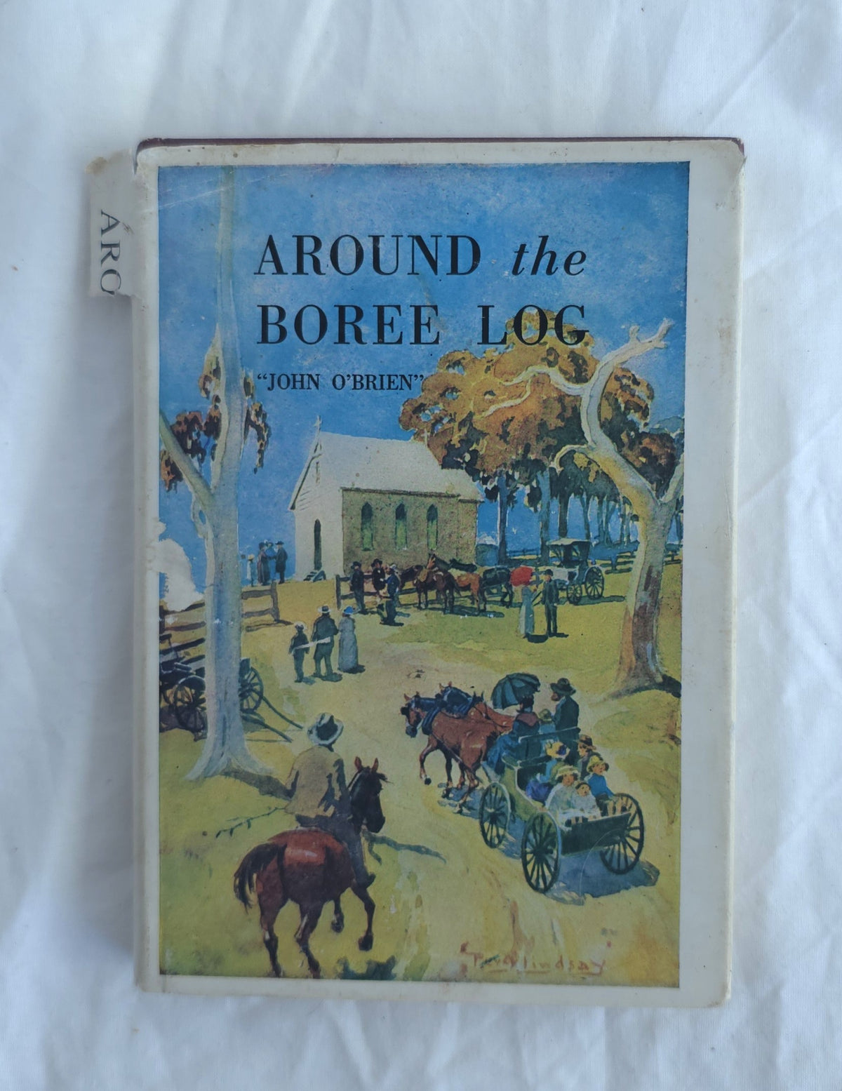 Vintage hardcover book titled 'Around the Boree Log' by John O'Brien, featuring a colorful illustration on the cover of a rural scene with people, horses, and a church, photographed on a white fabric background.