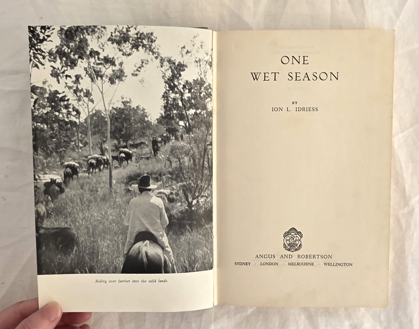 Open book titled 'One Wet Season' by Ion L. Idriess, showing a black and white photo of people on horseback in a field.