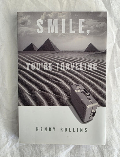 Book cover with pyramids and text 'SMILE, YOU'RE TRAVELING' by Henry Rollins on a textured white background.