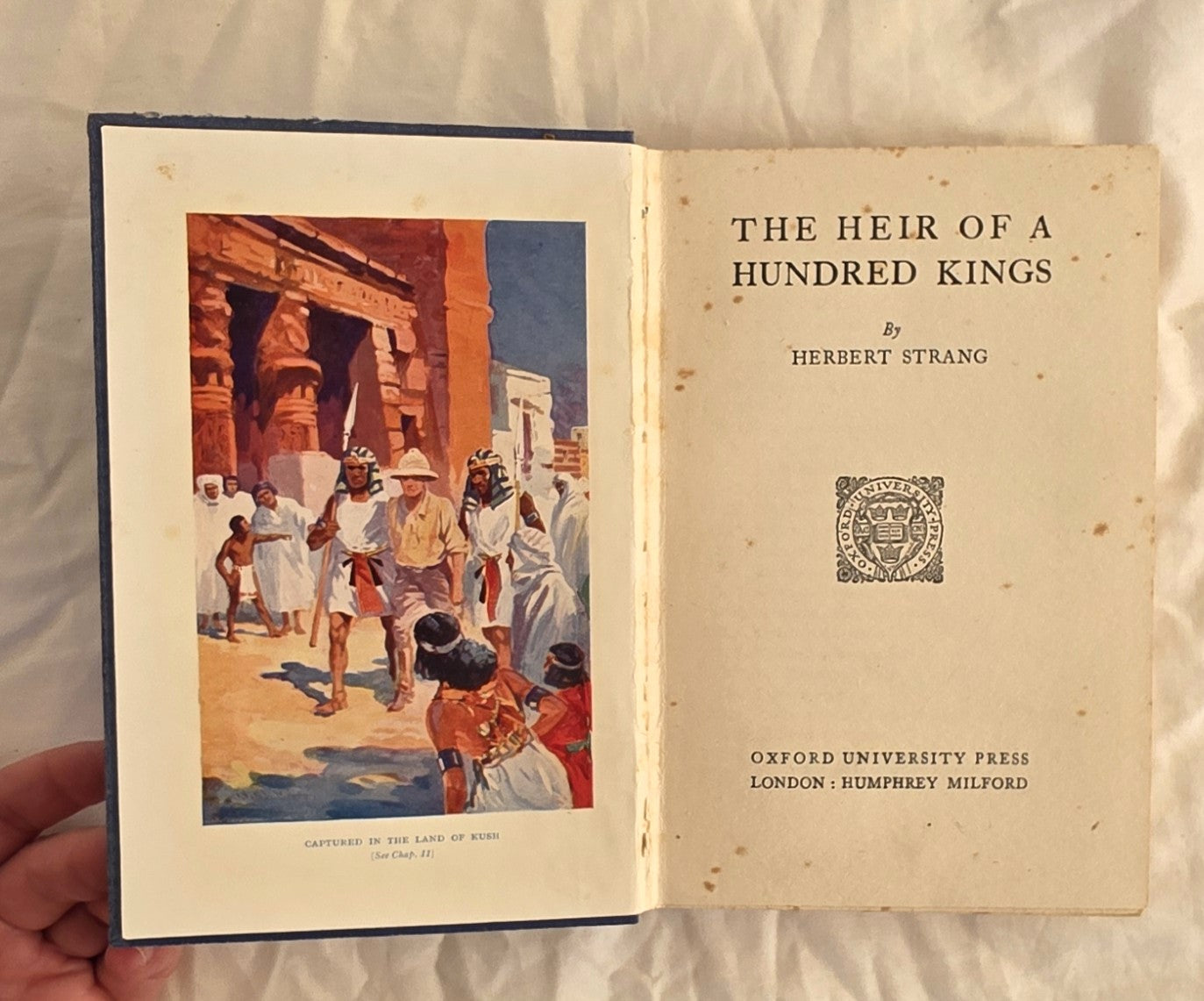 Open book titled 'The Heir of a Hundred Kings' by Herbert Strang, held against a neutral background.