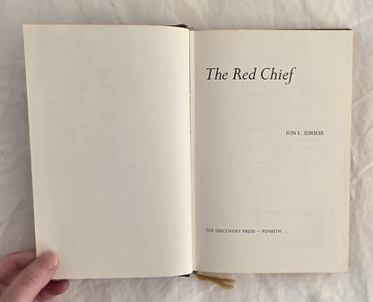Open book titled 'The Red Chief' by Ion L. Idriess held by a hand on a textured surface.